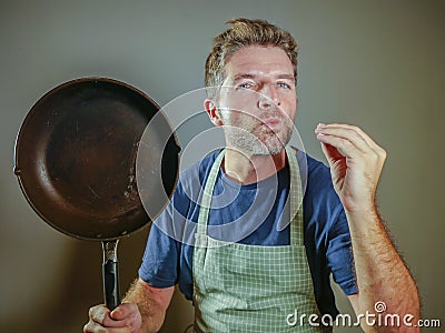 Young happy and attractive home cook man holding cooking pan smiling satisfied and proud giving delicious hand fingers sign as dom Stock Photo