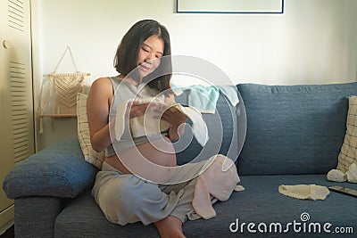 Young happy and beautiful Asian Korean woman pregnant on couch enjoying playful with little clothes of upcoming baby smiling Stock Photo