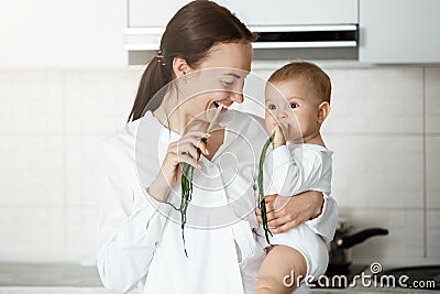 Young happy beautiful mother and little cute baby boy with brown eyes smiling and eating green onion in the kitchen for Stock Photo