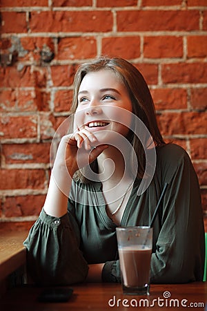 Young happy blond woman in green blouse sitting near window against red brick wall at the cafe with c cocoa glass Stock Photo