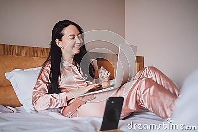 Young happy brunette woman with wireless earphones lying in bed in pink silk pajamas with smartphone and laptop computer Stock Photo