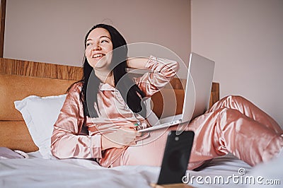 Young happy brunette woman with wireless earphones lying in bed in pink silk pajamas with smartphone and laptop computer Stock Photo