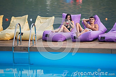 Young happy couple enjoying with drinks on cushioned loungers by swimming pool on the background of river Stock Photo