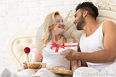 Young Happy Couple Lying In Bed, Hispanic Man Give Woman Surprise Present Envelope With Ribbon, Anniversary Celebration Stock Photo