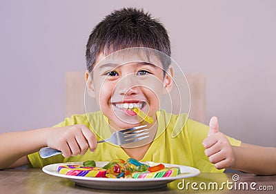 Young happy and excited male kid smiling cheerful eating dish full of candy and lollipop sitting at table isolated on grey Stock Photo