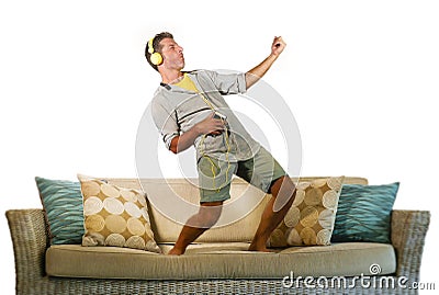 Young happy and excited man jumping on sofa couch listening to music with mobile phone and headphones playing air guitar crazy hav Stock Photo