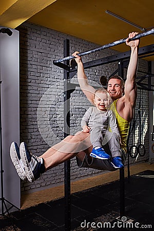 Young happy father doing pull ups on the bar with son on his legs at the cross fit gym against brick wall. Stock Photo