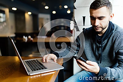 Young happy man smiling and holding a smartphone sitting with laptop in cafe. Portrait man reading a message with smartphone in ca Stock Photo