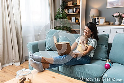Young happy mother reading a fairytale story book to her baby. Mommy and kid sitting on sofa at home enjoying in imagination. Stock Photo