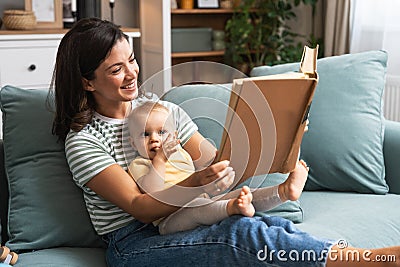 Young happy mother reading a fairytale story book to her baby. Mommy and kid sitting on sofa at home enjoying in imagination. Stock Photo