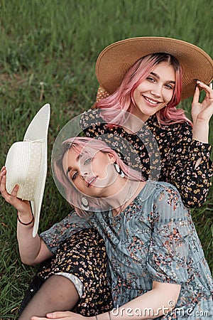 Young happy pink-haired woman in black stylish dress in straw hat sits with beautiful girlfriend with pink hair in blue summer Stock Photo