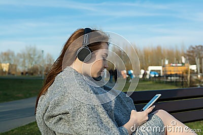 Young, happy redhead girl in the spring in the park near the river listens to music through wireless bluetooth headphones Stock Photo