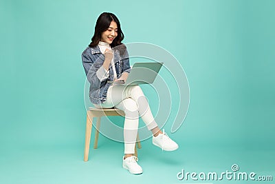 Young happy smiling Asian woman holding laptop computer and sitting on chair on green background. Stock Photo