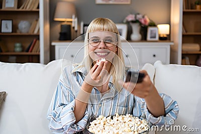 Young happy woman changing channels with remote control while watching TV and eating popcorn in the evening at home. Relaxed Stock Photo