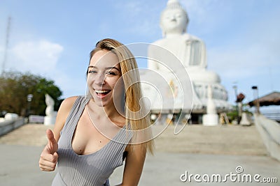 Young happy woman showing thumbs up, white Buddha statue in Phuket in background. Stock Photo