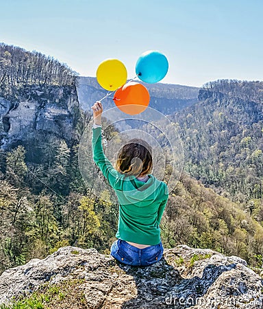 Young happy woman sitting on the edge of the cliff holding colorful balloons in her hands Stock Photo