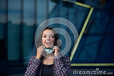 Happy woman takes off protective medical mask outdoors against modern city background. Pandemic Covid-19 is over Stock Photo