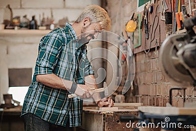 Attractive blonde young hipster bearded man by profession carpenter builder nailing wood board with hammer on a wooden Stock Photo