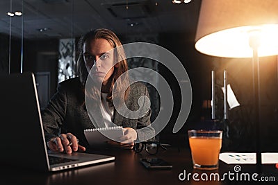 Young hipster businessman sits by the wooden table with lamp, laptop and smartphone at night office. Man with long hair work with Stock Photo