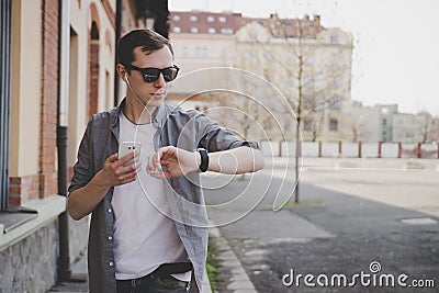 Young hipster man walking on the street, looking at watch and using his smartphone. With copy space Stock Photo