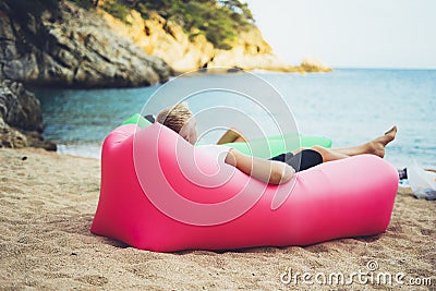 Young hipster relaxing on coastline beach on inflatable lazy air pouffe sofa, person tourist enjoy sunny day on background coast Stock Photo