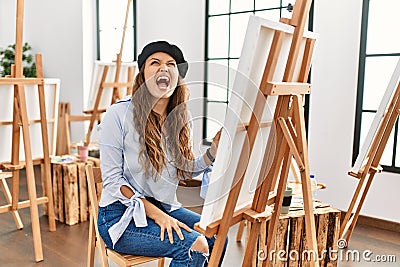 Young hispanic artist woman painting on canvas at art studio angry and mad screaming frustrated and furious, shouting with anger Stock Photo