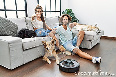 Young hispanic couple relaxing at home by vacuum robot with angry face, negative sign showing dislike with thumbs down, rejection Stock Photo