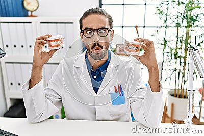 Young hispanic dentist man holding denture with orthodontic braces making fish face with mouth and squinting eyes, crazy and Stock Photo