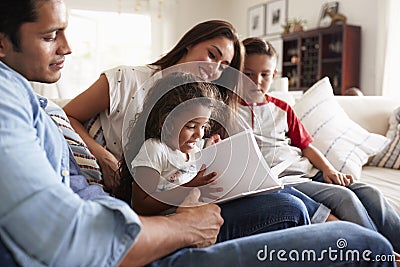 Young Hispanic family of four sitting on the sofa reading book together in their living room Stock Photo