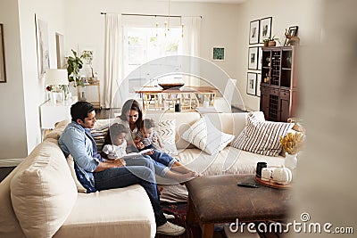 Young Hispanic family sitting on sofa reading a book together in the living room, seen from doorway Stock Photo