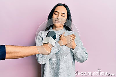 Young hispanic girl being interviewed by reporter holding microphone smiling with hands on chest, eyes closed with grateful Stock Photo