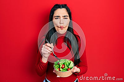 Young hispanic girl eating salad making fish face with mouth and squinting eyes, crazy and comical Stock Photo