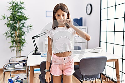 Young hispanic girl standing at pediatrician clinic mouth and lips shut as zip with fingers Stock Photo