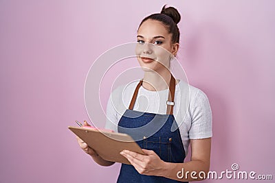 Young hispanic girl wearing professional waitress apron taking order smiling with a happy and cool smile on face Stock Photo