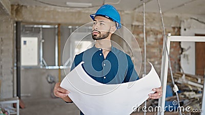 Young hispanic man architect wearing hardhat looking at blueprints at construction site Stock Photo