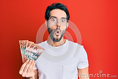 Young hispanic man holding australian dollars scared and amazed with open mouth for surprise, disbelief face Stock Photo