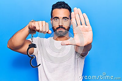 Young hispanic man holding prisoner handcuffs with open hand doing stop sign with serious and confident expression, defense Stock Photo
