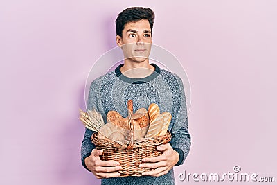 Young hispanic man holding wicker basket with bread smiling looking to the side and staring away thinking Stock Photo