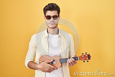 Young hispanic man playing ukulele over yellow background relaxed with serious expression on face Stock Photo