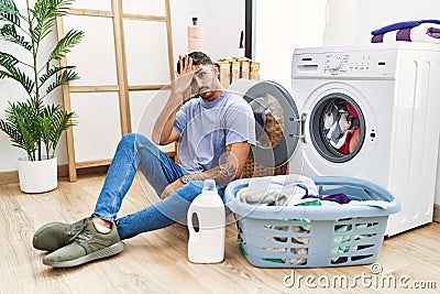 Young hispanic man putting dirty laundry into washing machine surprised with hand on head for mistake, remember error Stock Photo