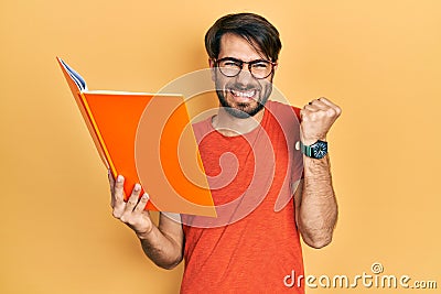 Young hispanic man reading book screaming proud, celebrating victory and success very excited with raised arm Stock Photo