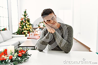 Young hispanic man sitting on the table by christmas tree sleeping tired dreaming and posing with hands together while smiling Stock Photo