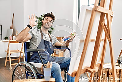 Young hispanic man sitting on wheelchair painting at art studio waiving saying hello happy and smiling, friendly welcome gesture Stock Photo