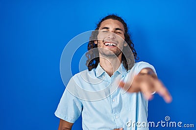 Young hispanic man standing over blue background laughing at you, pointing finger to the camera with hand over body, shame Stock Photo