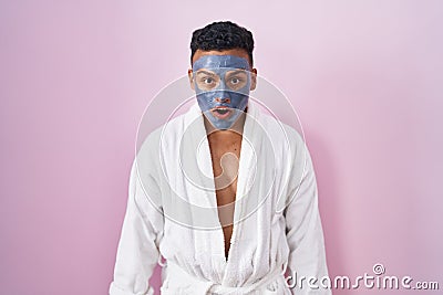 Young hispanic man wearing beauty face mask and bath robe afraid and shocked with surprise and amazed expression, fear and excited Stock Photo
