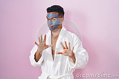 Young hispanic man wearing beauty face mask and bath robe afraid and terrified with fear expression stop gesture with hands, Stock Photo
