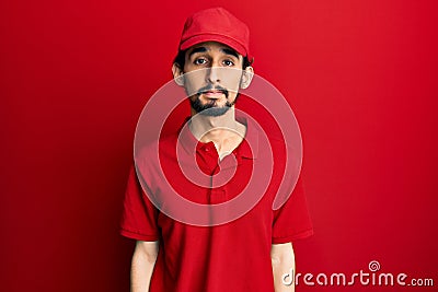 Young hispanic man wearing delivery uniform and cap with serious expression on face Stock Photo