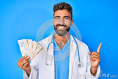 Young hispanic man wearing doctor uniform holding uk pounds banknotes smiling happy pointing with hand and finger to the side Editorial Stock Photo