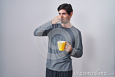 Young hispanic man wearing pajama drinking a cup of coffee smelling something stinky and disgusting, intolerable smell, holding Stock Photo