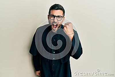 Young hispanic man wearing priest uniform standing over white background angry and mad raising fist frustrated and furious while Stock Photo